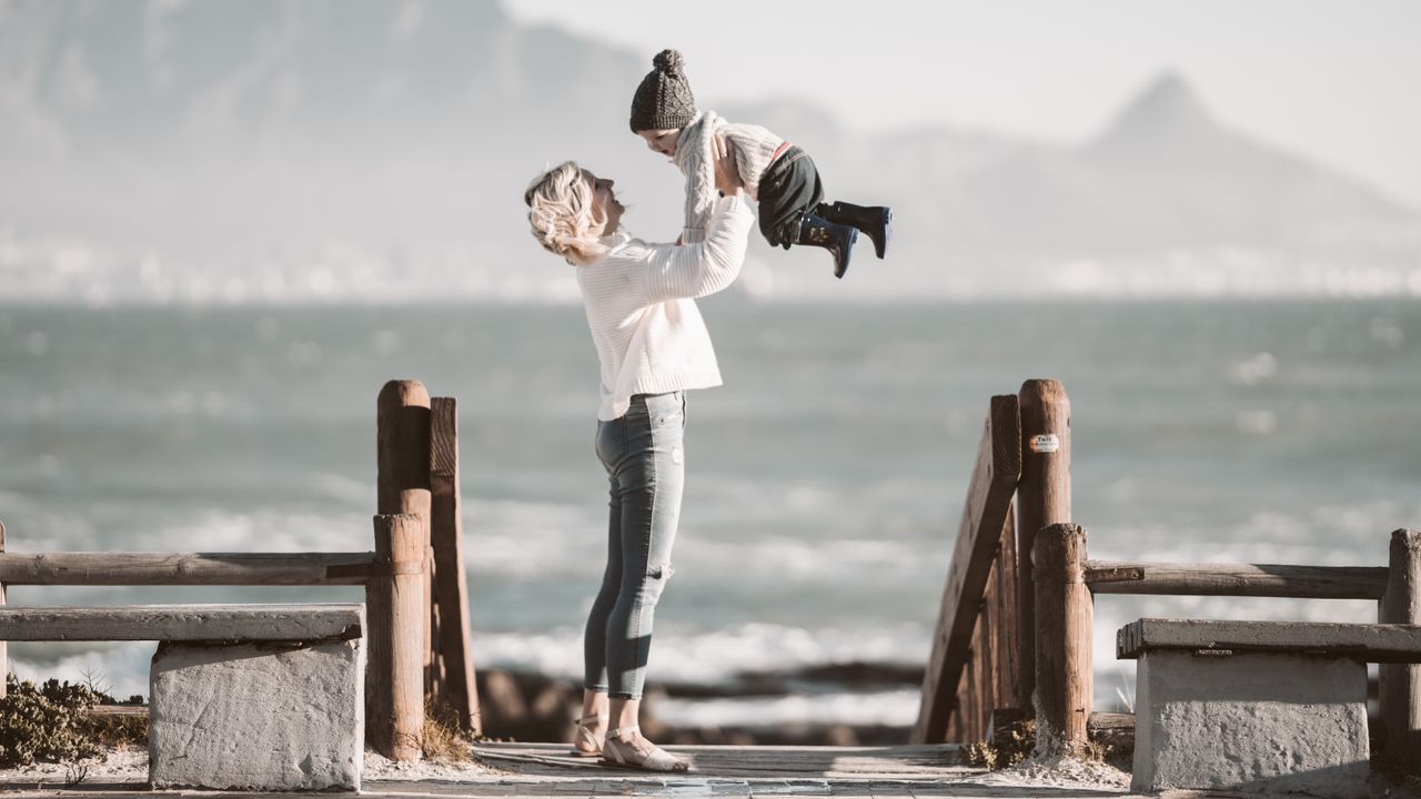 A joyful mother lifting her child near the beach on a sunny day.